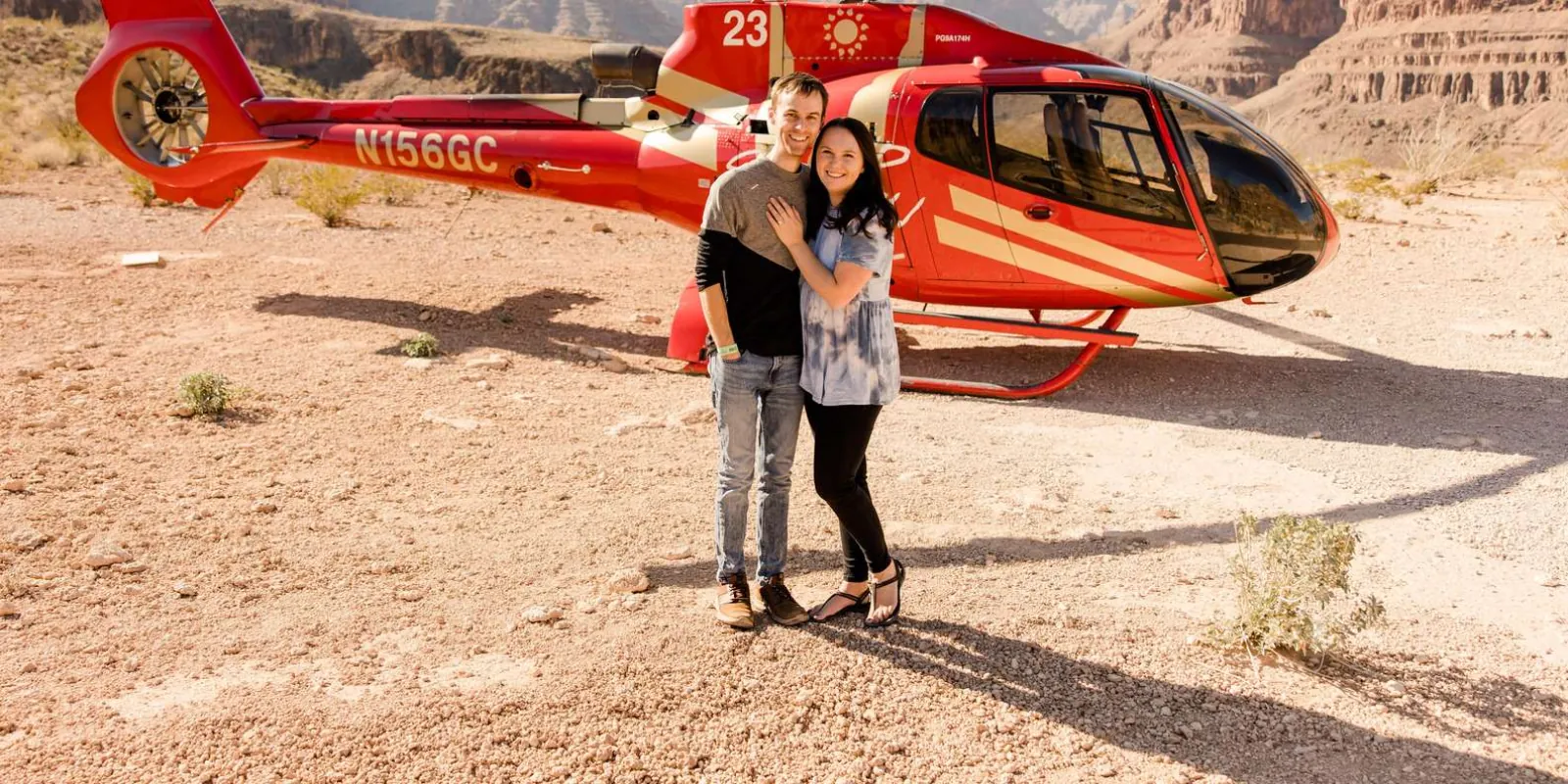 Two guests hugging beside a red Papillon EcoStar helicopter parked on the Grand Canyon floor
